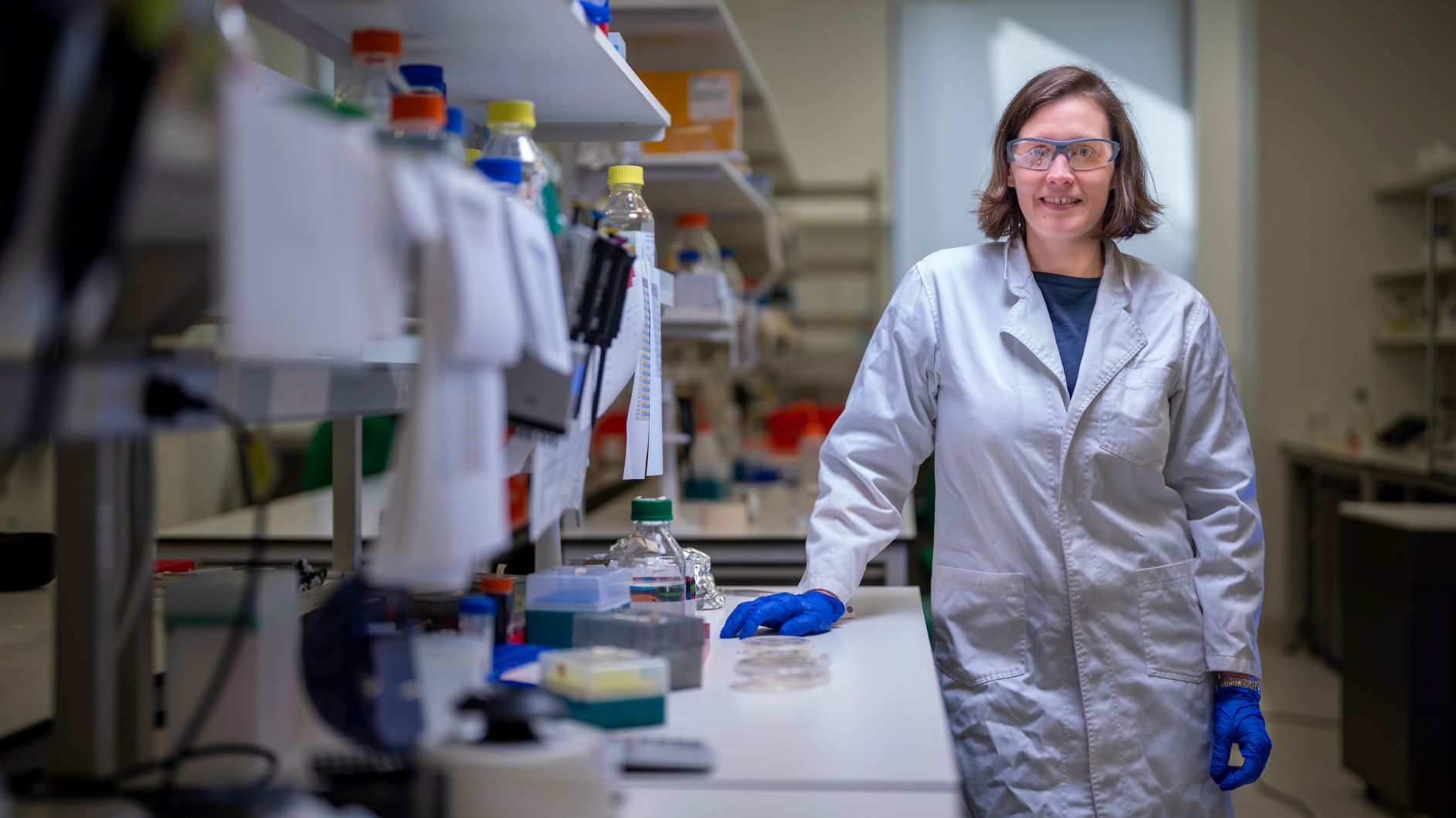 A woman in a lab wearing a lab coat and safety glasses