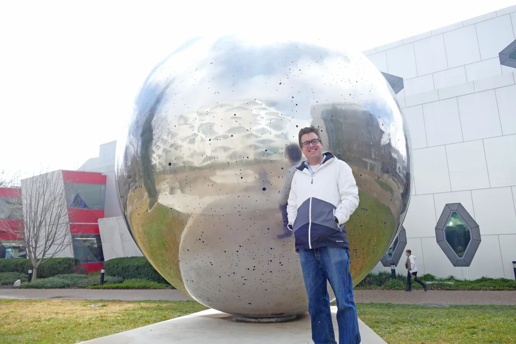 A man is standing in front of a large spherical sculpture on a university campus.