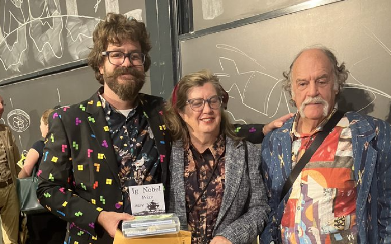 A man in a brightly coloured blazer is holding an Ig Nobel Prize trophy. He is joined by his two parents.