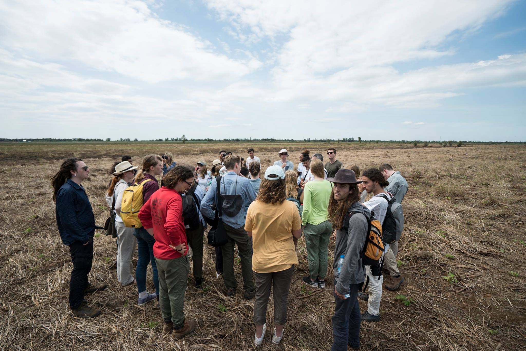 A group of people in casual clothing are gathered in a field under a cloudy sky, attentively listening to a speaker.