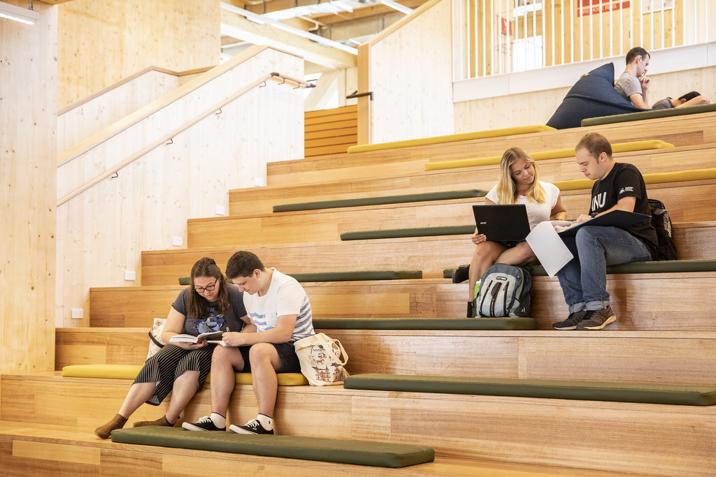 Students study and use laptops on the wooden bleachers inside a modern, well-lit building.