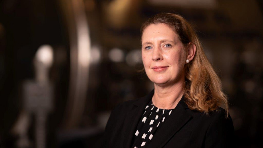A woman with light brown hair, wearing a black blazer and a white blouse with a geometric pattern, smiles softly while posing for a portrait in a dimly lit room with blurred metallic sculptures in the background.