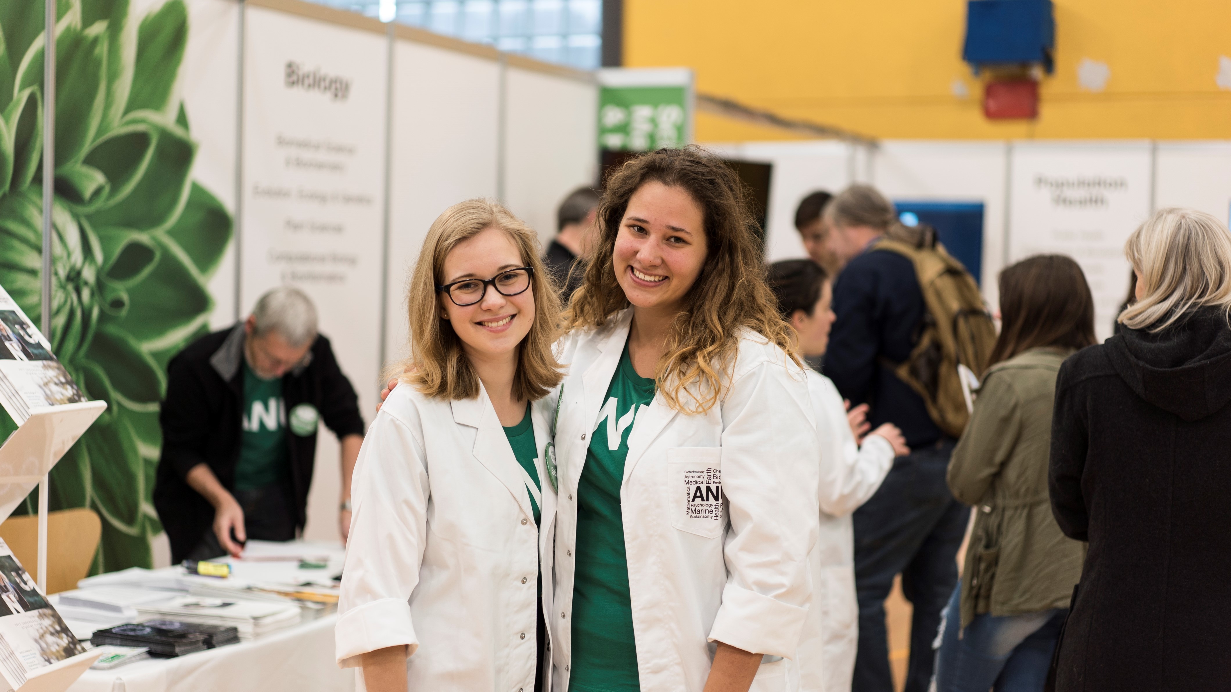 Two smiling women in white lab coats standing at an informational booth at a career fair.