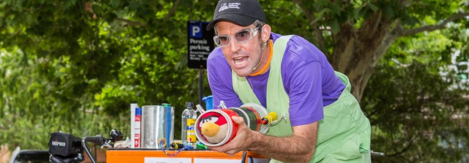 A man in a purple and green apron works on a project at a cluttered table, surrounded by cans and tools.