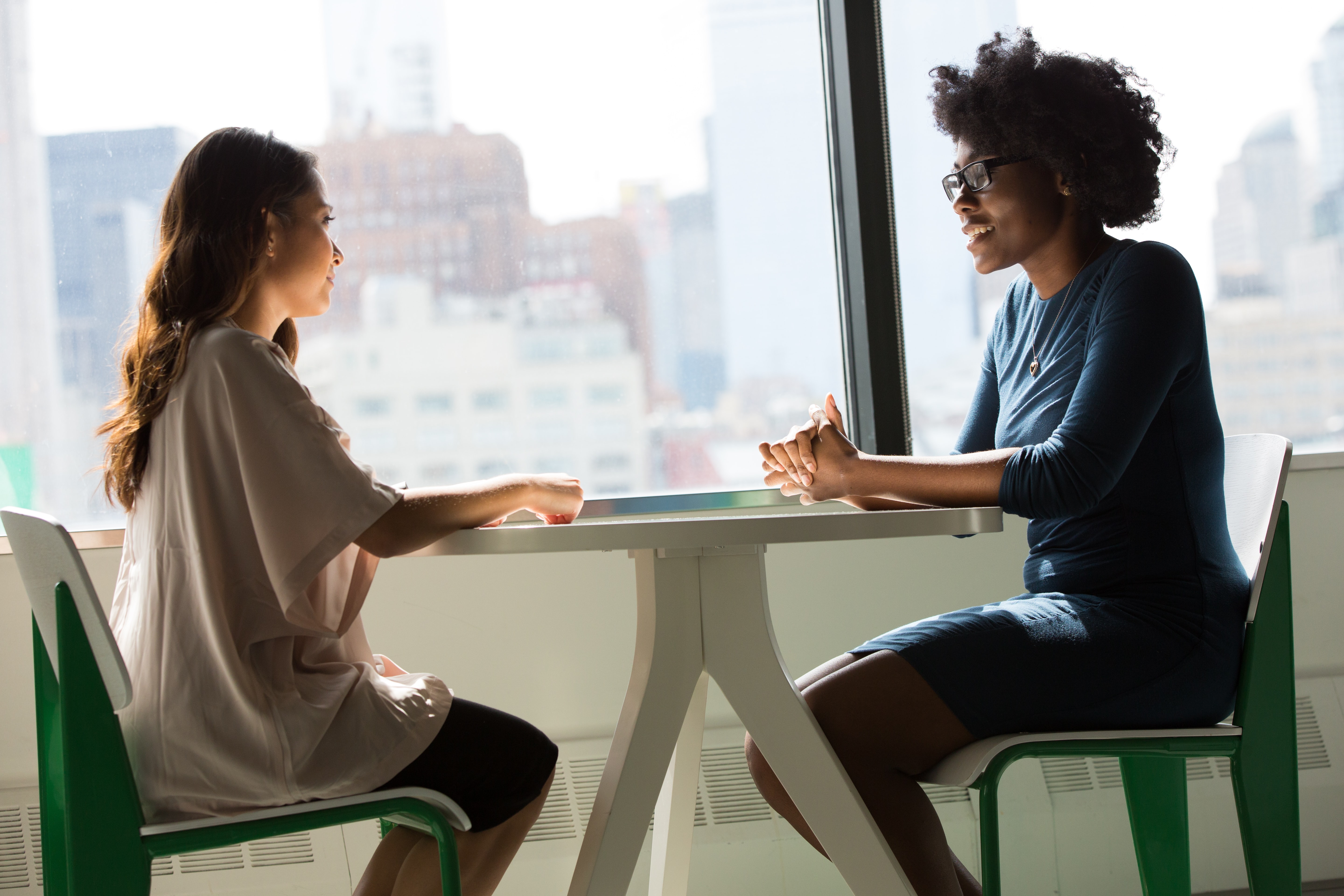 Two women are seated at a table by a large window in a well-lit room, engaged in conversation.
