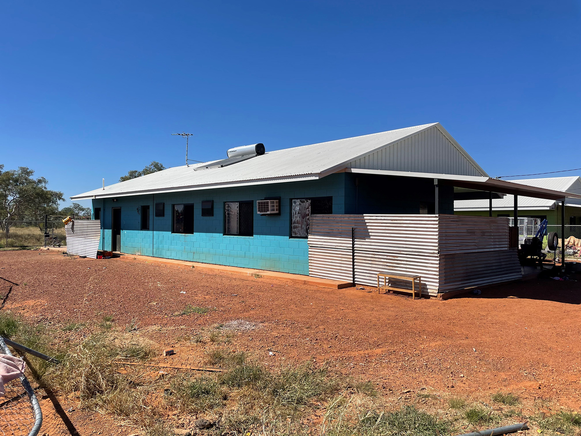 A single-story light blue house with a white roof, surrounded by a red dirt yard under a clear blue sky.