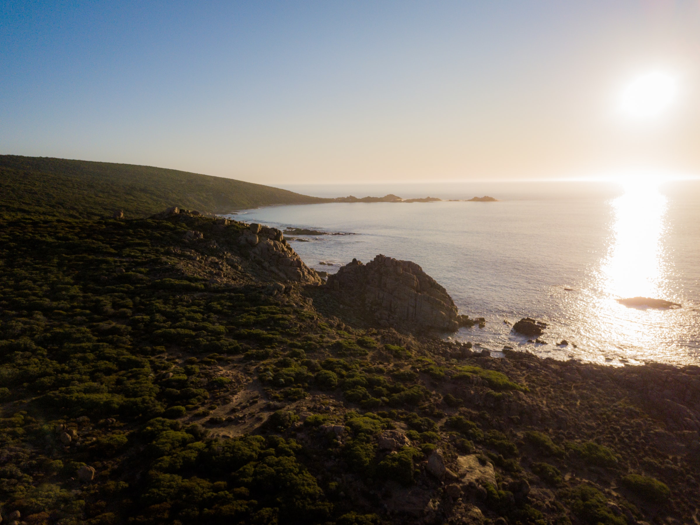Aerial view of a rugged coastline with green vegetation and rocky outcrops, overlooking a sunlit ocean during sunset.