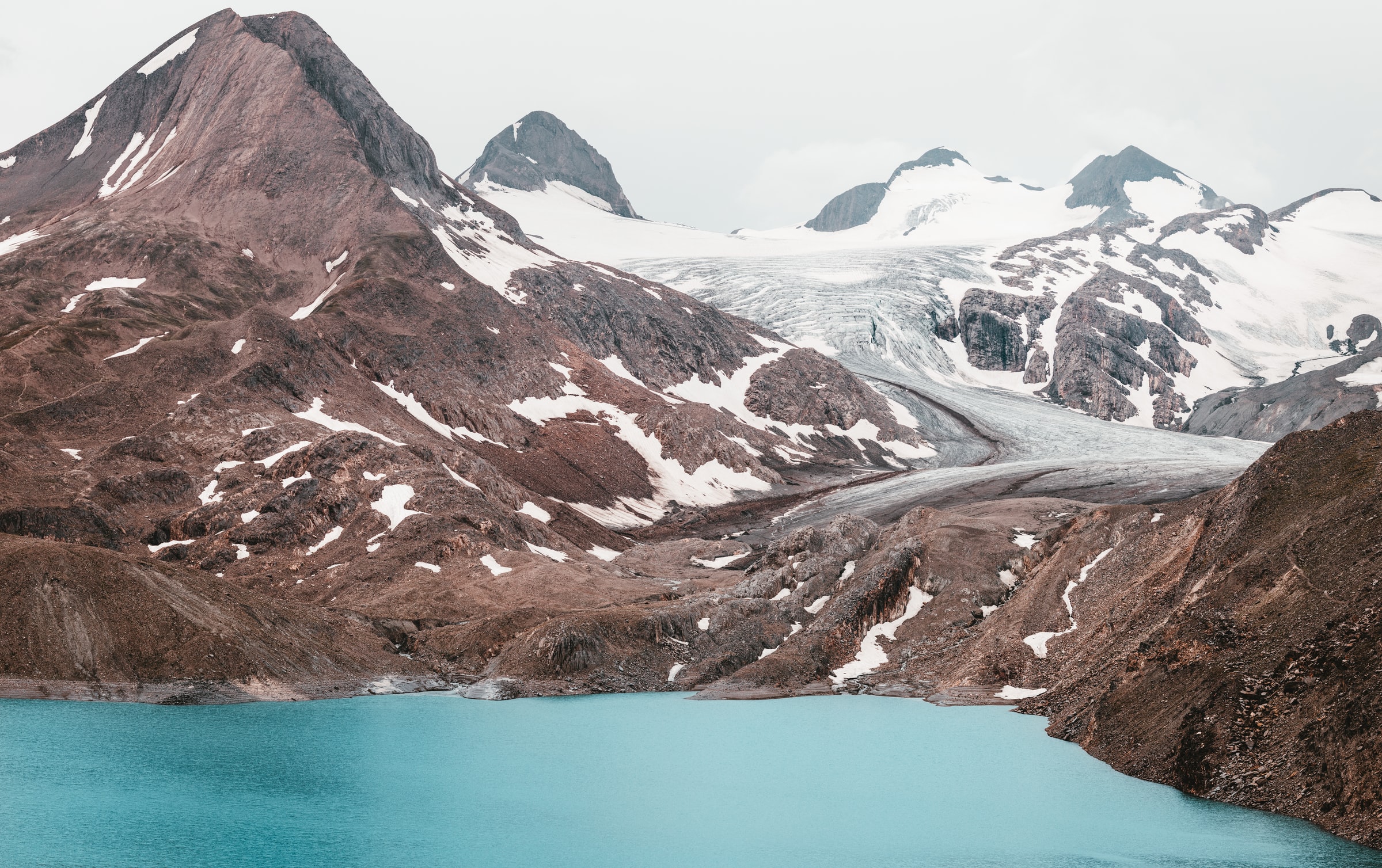 A glacial mountain landscape with a vivid turquoise lake in the foreground surrounded by rugged brown and snowy peaks.