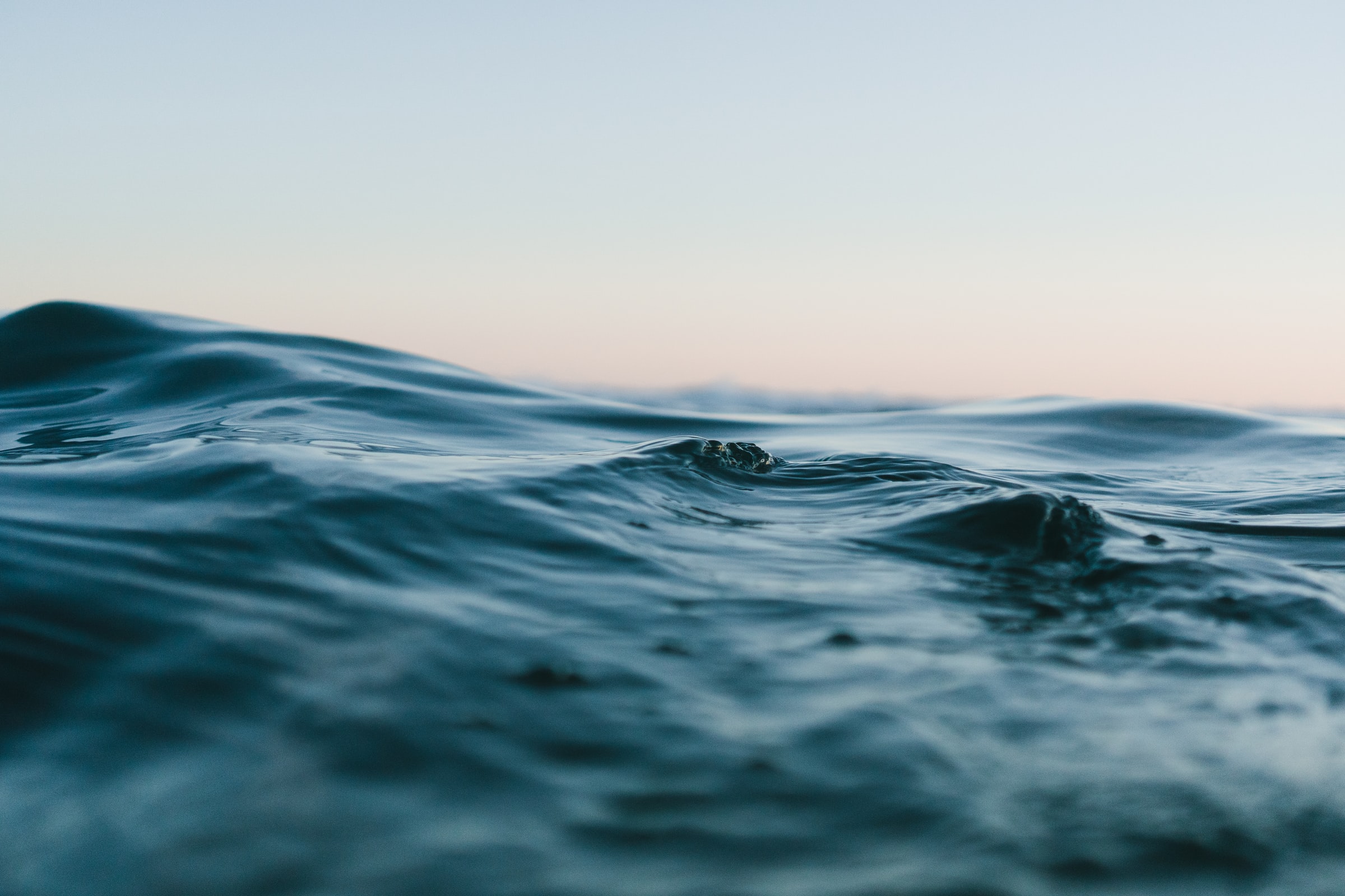 Close-up photo of calm ocean water with gentle waves and a soft blue sky in the background.