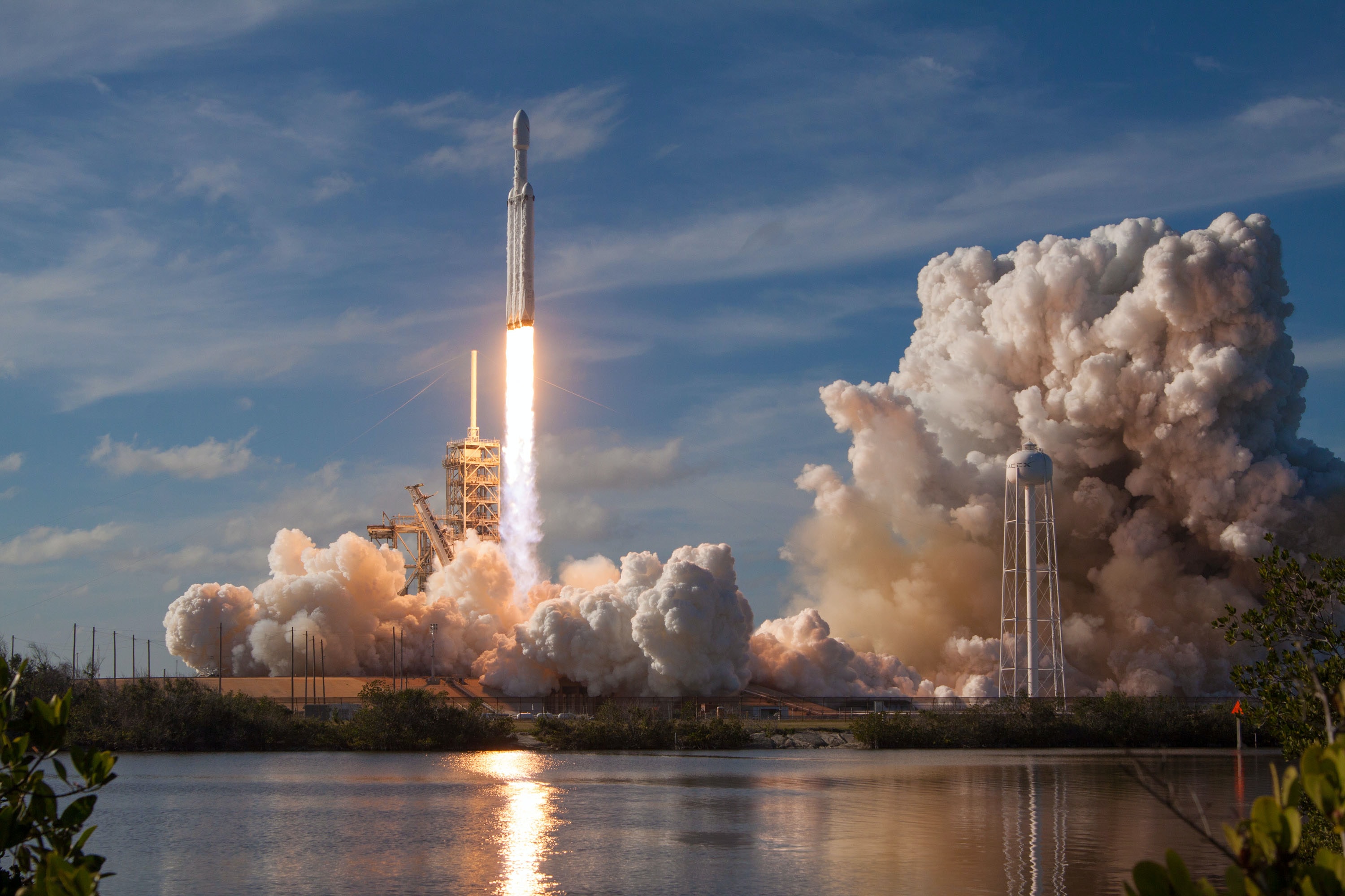 A rocket launches from a launchpad, emitting a powerful plume of smoke and fire, with water in the foreground reflecting the scene.