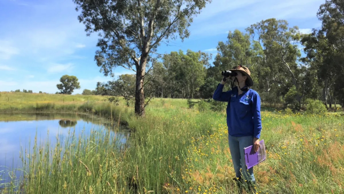 A researcher holds binoculars while looking across a field. A lake is visible in the background.