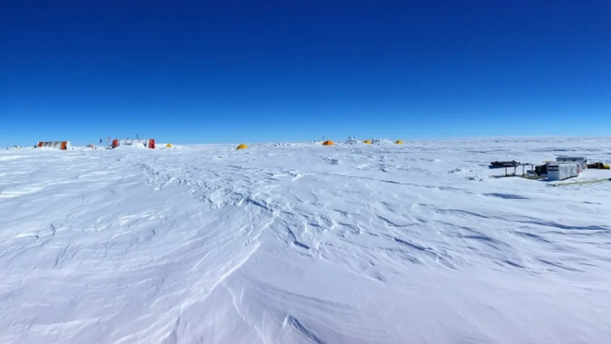 An ice sheet is covered in white snow under a blue sky, there are small huts in the background.