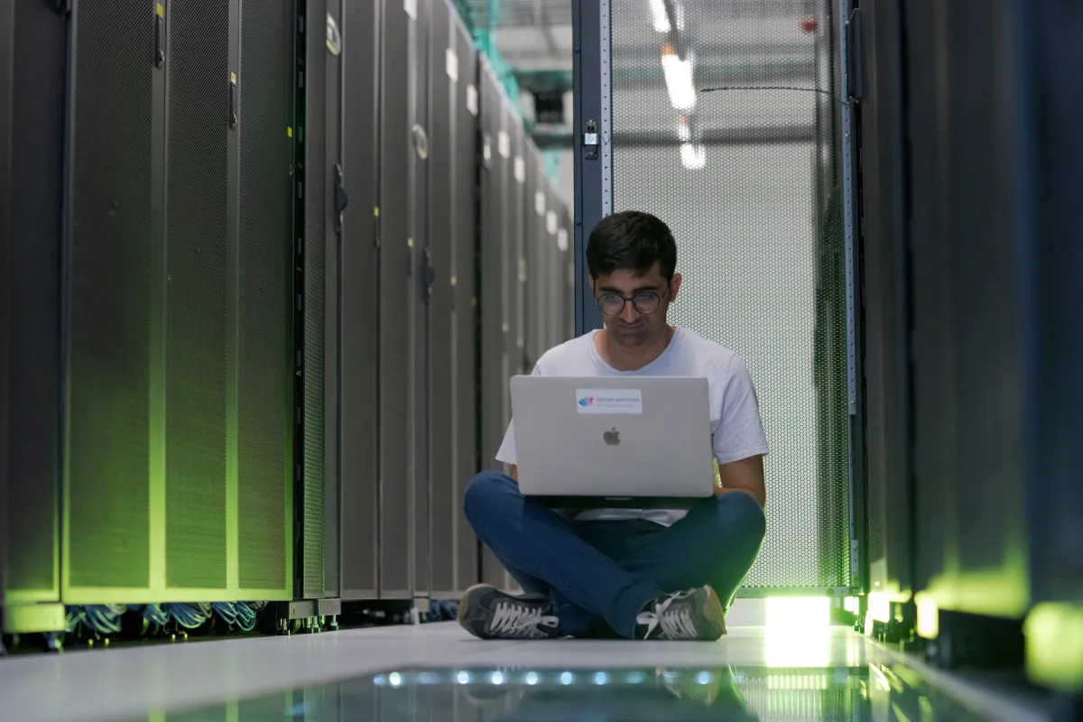 A man sitting in a computer server room, looking at a laptop