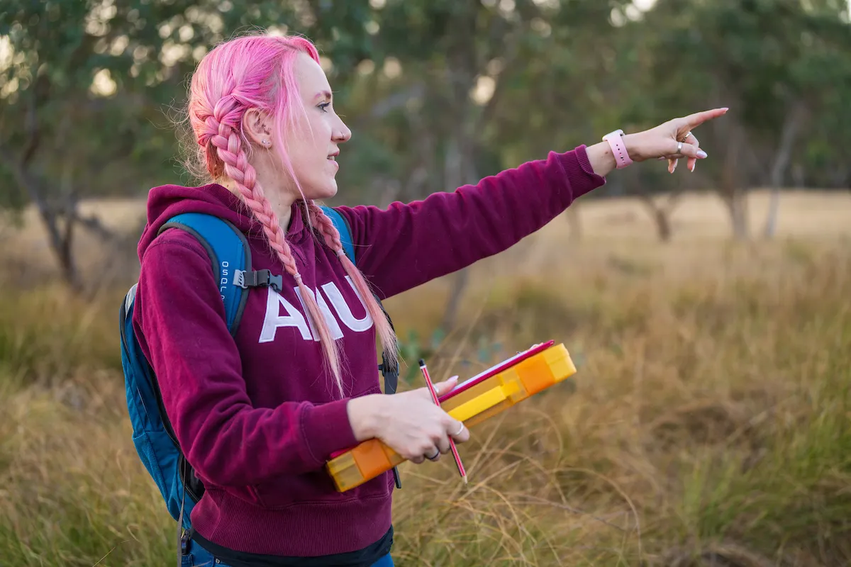 A woman with pink hair and holding a clipboard pointing to something