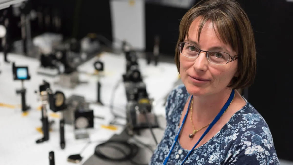 A women looks to the camera. She is standing next to optical space laboratory equipment.
