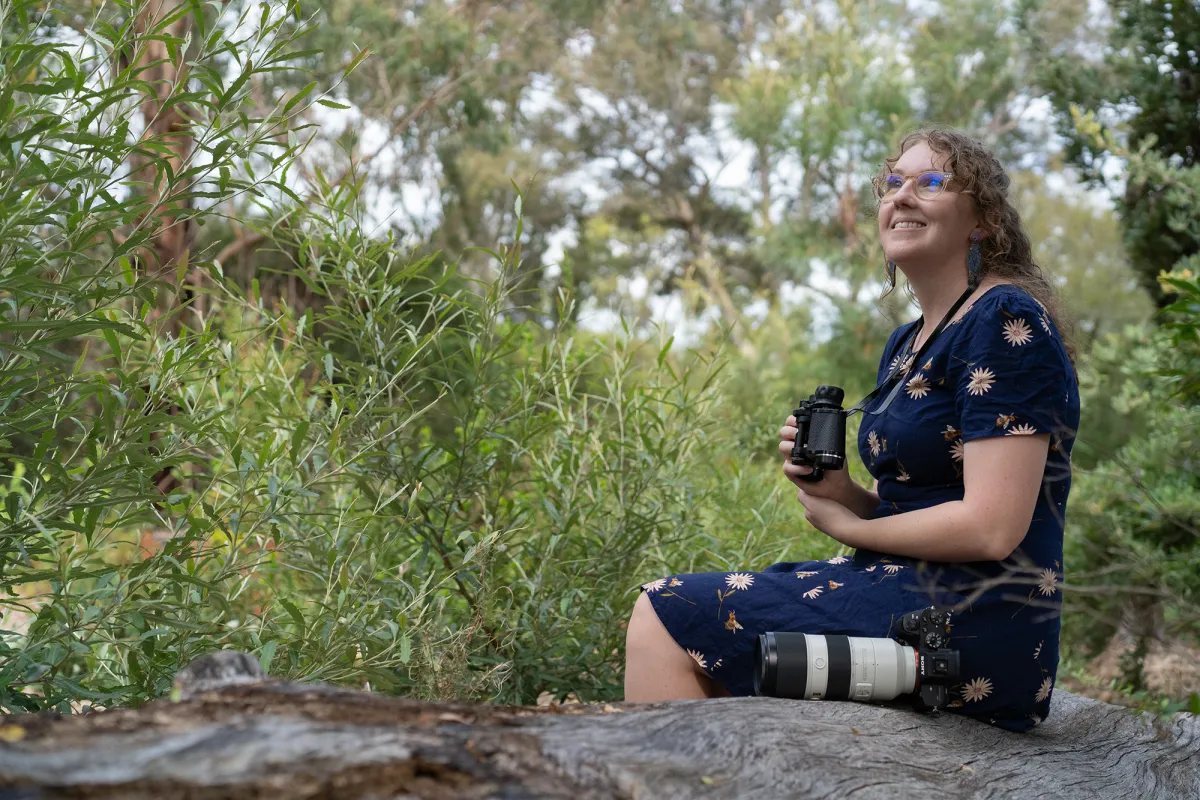 Woman holds binoculars witha camera on a log