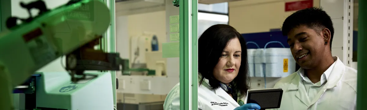 A woman and man are both wearing lab coats and standing in front of scientific equipment