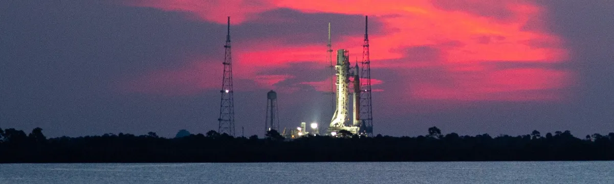 A rocket on a launchpad near water against a sunset