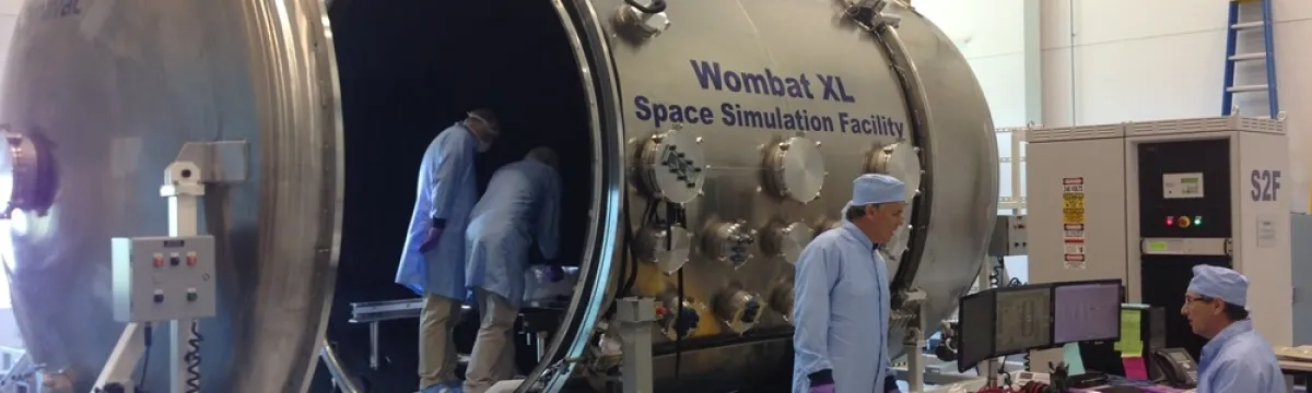 Technicians work in a lab around the Wombat XL Space Simulation Facility, a large metallic chamber with an open hatch.