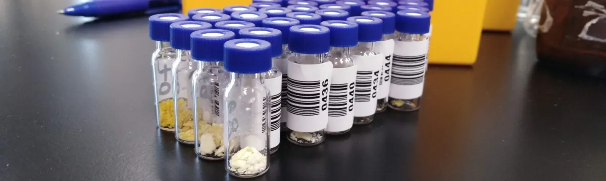 Multiple small sample bottles with blue caps, containing various substances, are lined up on a laboratory bench.