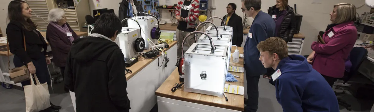 A group of people attentively watching a demonstration of 3D printers in a workshop setting.