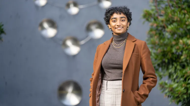 Mallika Sinha outside in front of a grey sculpture. Mallika is wearing a brown coat and is smiling towards the camera