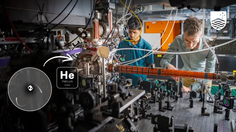 Yogesh Sridhar and Sean Hodgman are working in a physics laboratory. There are lots of cords and metal equipment. With a helium atom graphic in the bottom left.