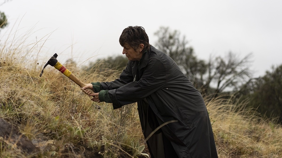 Lady in large raincoat, using mattock that looks like a large pickaxe to remove weeds from an embankment.
