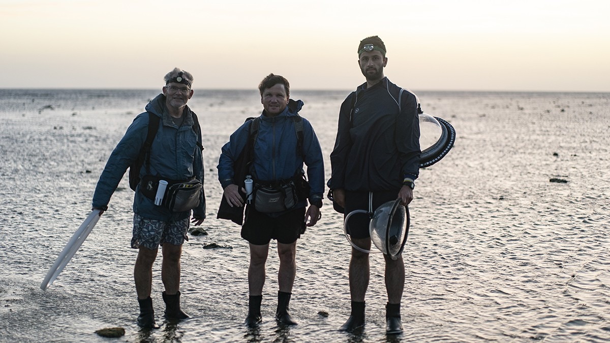 Three researchers face the camera on the reef flat at sunset, ready to head back to the island with their gear.