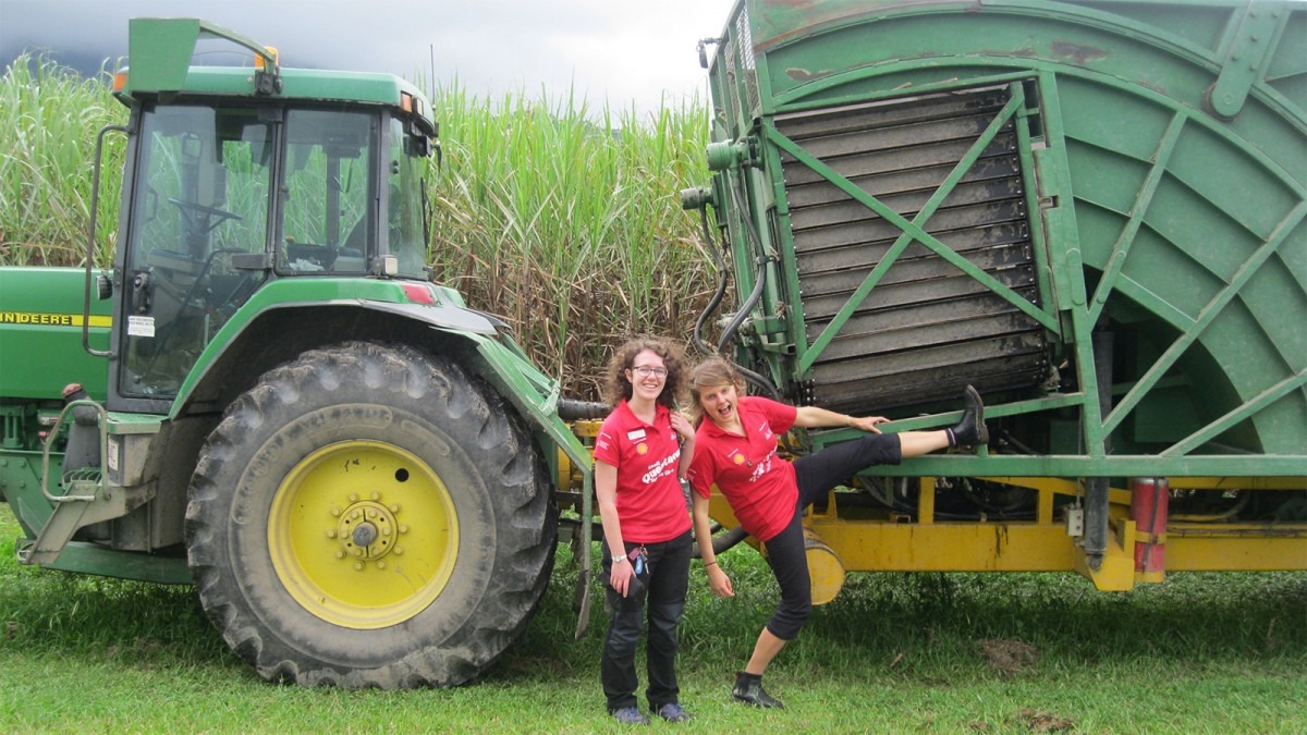 Two people wearing Questacon Science Circus shirts stand in front of a tractor in a cane field.