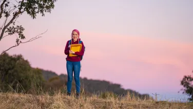 A woman with pink hair and holding a clipboard standing in some bushy grass