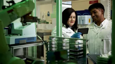 A woman and man are both wearing lab coats and standing in front of scientific equipment
