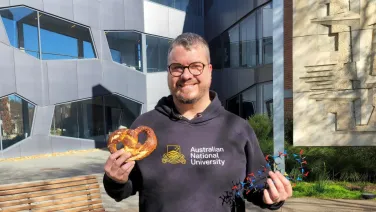 A researcher holds up a large pretzel in one hand and a 3D molecule model in the other. He is smiling at the camera. Behind him is the Research School of Chemistry building.