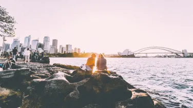 People sitting on some rocks by the water side at dusk. The Sydney Harbour Bridge is in the background.