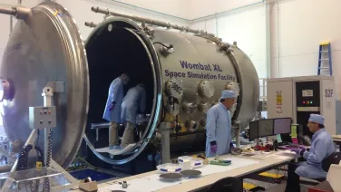 Technicians work in a lab around the Wombat XL Space Simulation Facility, a large metallic chamber with an open hatch.