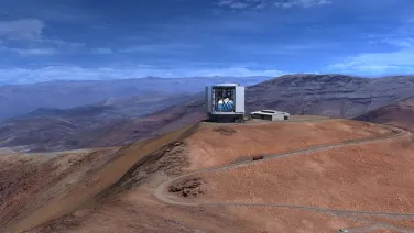 A large astronomical observatory sits atop a barren, dusty mountain under a clear blue sky.