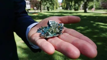 Close up of hand holding the heavy metal element bismuth, showing a crystal structure and iridescent surface.
