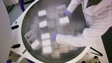 A scientist wearing gloves places containers inside a large, circular laboratory machine.