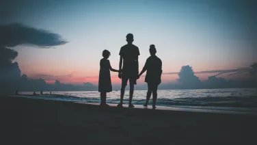 Three people stand hand-in-hand on a beach at dusk, silhouetted against a vibrant sunset and ocean waves.