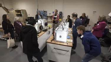 A group of people attentively watching a demonstration of 3D printers in a workshop setting.