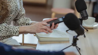 Close up of someone holding a smartphone, sitting at a table with microphones and a notebook in front of them.